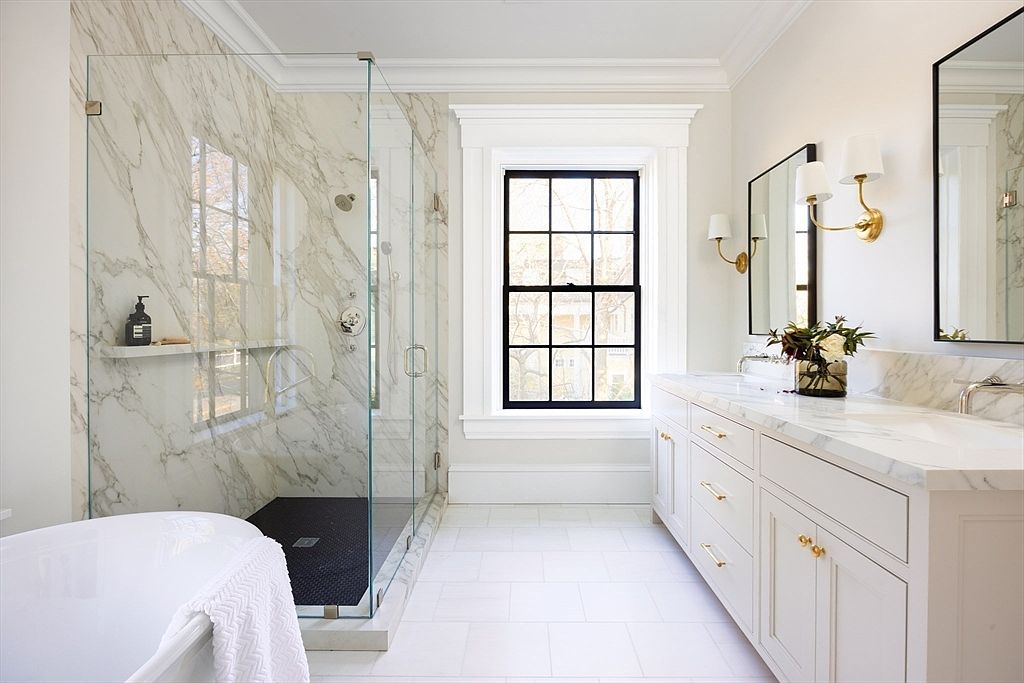 This luxurious primary bathroom features a spacious walk-in glass shower with marble-tiled walls and a dark stone floor, adjacent to a sleek white double vanity with marble countertops. A large, black-framed window serves as a striking focal point, allowing natural light to flood the room and highlighting the elegant gold hardware and sconce lighting. The overall aesthetic is a sophisticated blend of classic marble textures and modern, clean-lined design.