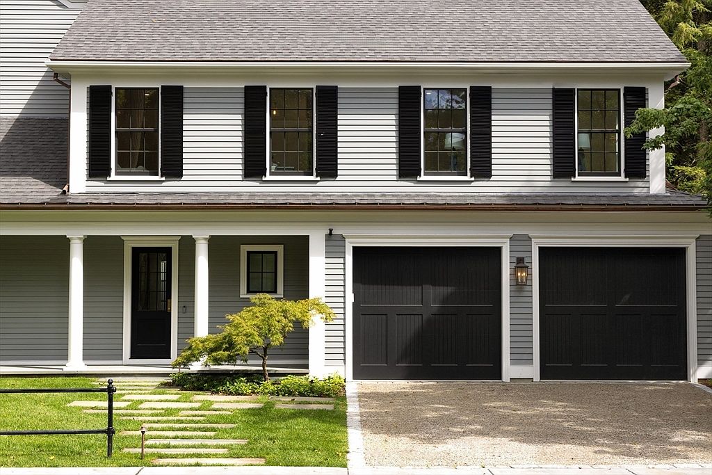 This charming two-story home features classic gray horizontal siding, crisp white trim, and striking black shutters and garage doors, creating a sophisticated and modern farmhouse aesthetic. The front exterior includes a welcoming covered porch with white columns, a stone paver walkway set into a manicured lawn, and a gravel driveway leading to a two-car garage. The perspective is a straight-on, eye-level shot that highlights the home's symmetrical design and curb appeal.