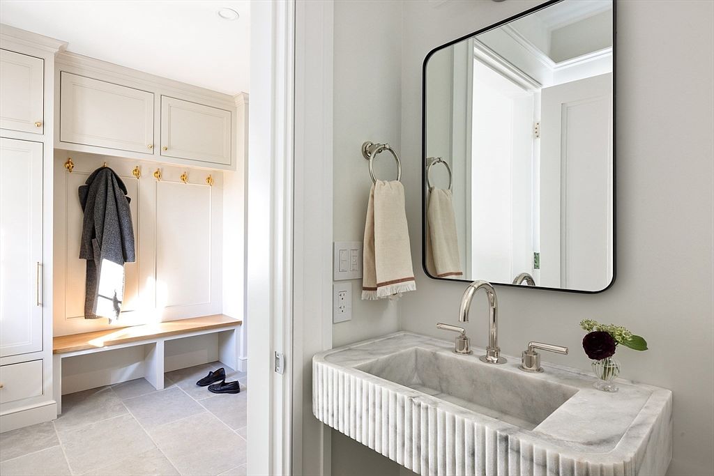 This elegant powder room features a striking fluted marble vanity sink paired with a sleek, black-framed mirror. Through the doorway, a bright mudroom area is visible, complete with built-in cabinetry, a wooden bench, and gold-toned hooks. The combination of high-end stone textures and functional storage creates a sophisticated and welcoming transition between spaces.