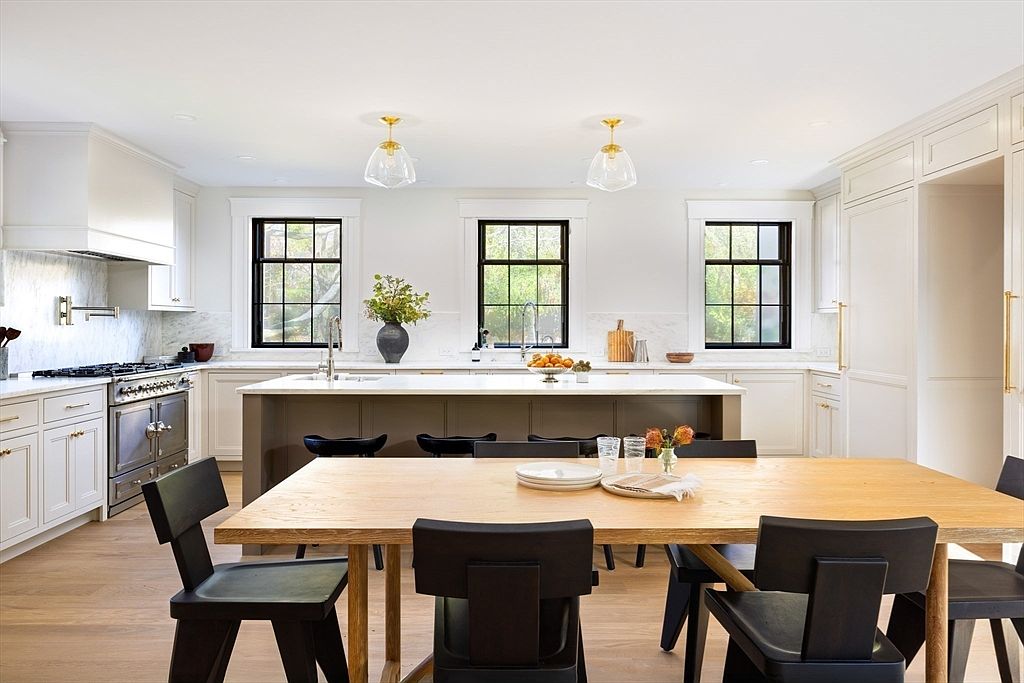 This bright and airy kitchen features a large central island with dark cabinetry, contrasting beautifully against the surrounding white perimeter cabinets and marble countertops. The space is anchored by a wooden dining table in the foreground, paired with modern black chairs, creating a seamless transition between cooking and dining areas. Large black-framed windows allow natural light to flood the room, highlighting the clean, sophisticated design and high-end finishes.
