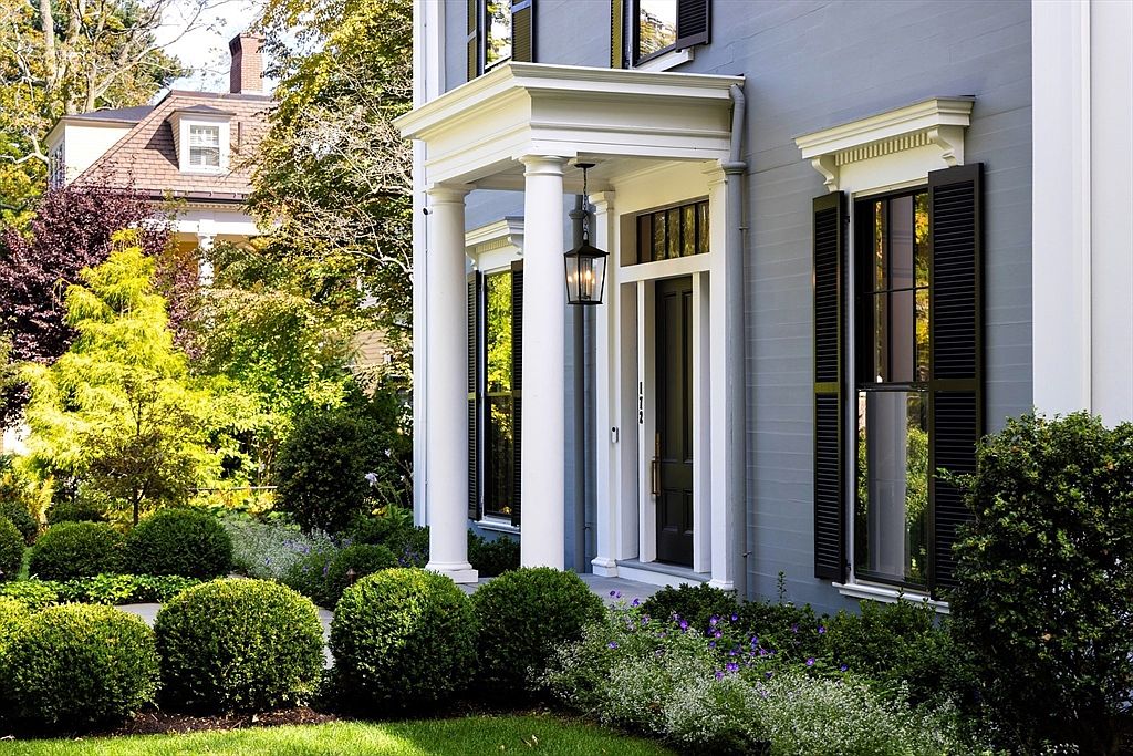 This elegant front entryway features a classic portico supported by two white columns, framing a dark-toned front door with a transom window. The home is finished in a sophisticated grey siding with black shutters, complemented by a meticulously landscaped garden of manicured boxwood shrubs and flowering perennials. The perspective is a ground-level eye view, capturing the inviting and well-maintained curb appeal of a traditional-style residence.