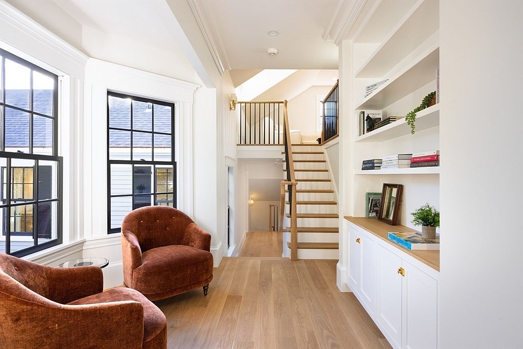 This bright and airy hallway features light oak hardwood flooring and a striking staircase with a wooden handrail and black metal balusters. To the left, two plush, rust-colored armchairs are positioned near large windows, while a built-in white bookshelf and cabinet unit provide elegant storage on the right. The perspective captures a clean, transitional design that seamlessly connects different levels of the home with a sense of warmth and sophistication.