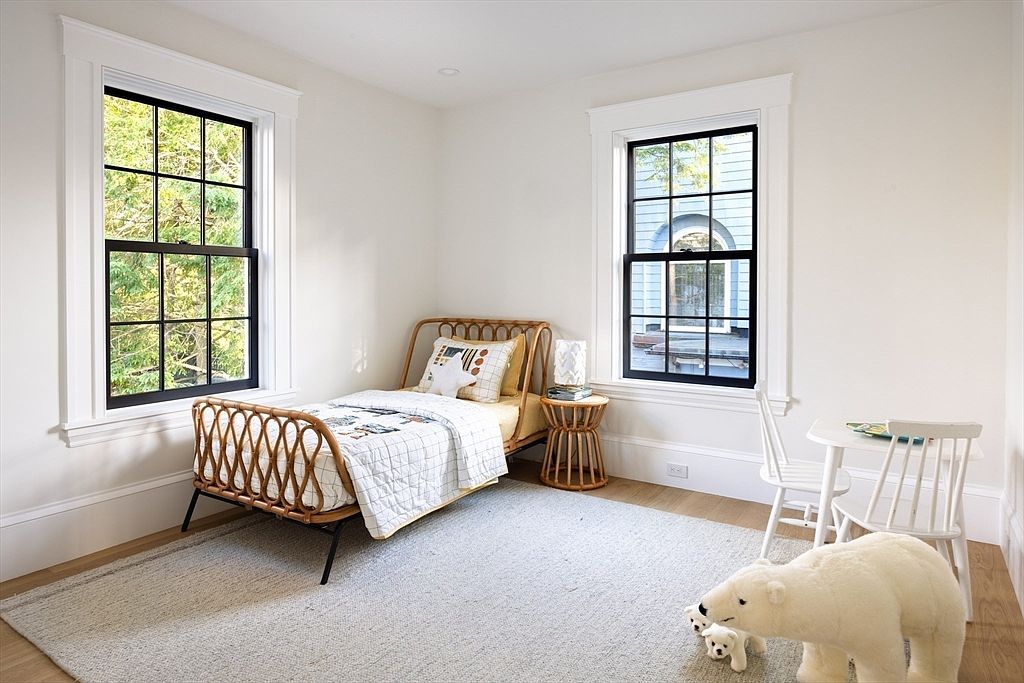 This charming, light-filled child's bedroom features a natural rattan bed frame and a matching side table, creating a warm and inviting aesthetic. The room is anchored by a large, neutral area rug and includes a small white table and chair set, perfect for play or study. Two large windows with black frames provide ample natural light, highlighting the clean white walls and minimalist decor.