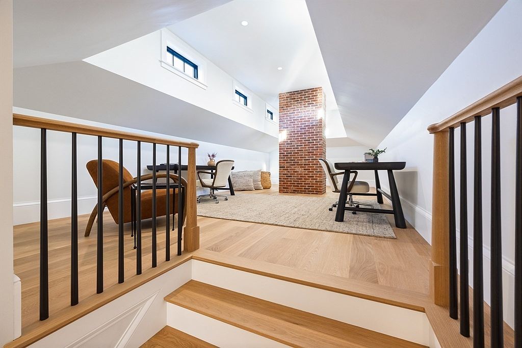 This bright and airy attic space has been converted into a functional home office, featuring light hardwood flooring and a prominent exposed brick chimney. The room is illuminated by natural light streaming through multiple skylights, creating a warm and inviting atmosphere. The perspective from the top of the stairs highlights the open-concept layout, showcasing a desk area and a comfortable seating nook.