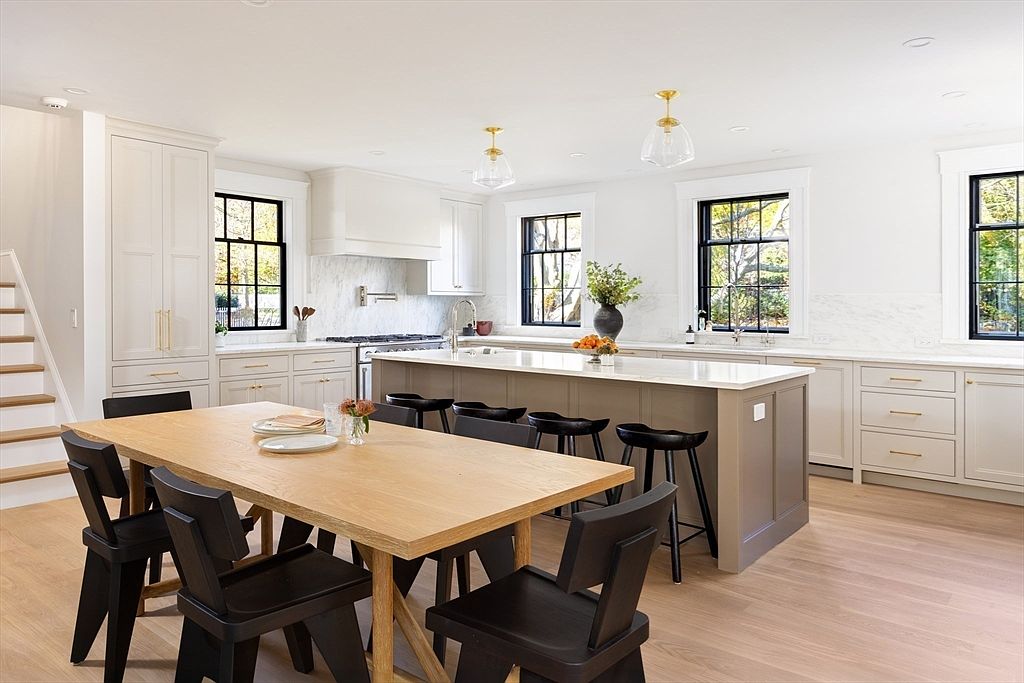 This bright and airy kitchen features a sophisticated blend of light-toned cabinetry and a contrasting dark-toned central island, complemented by warm wood flooring and a large dining table. The space is illuminated by elegant pendant lighting and natural light streaming through black-framed windows, creating a modern, high-end aesthetic. The open-concept layout seamlessly integrates the dining area with the gourmet kitchen, making it an ideal space for both cooking and entertaining.