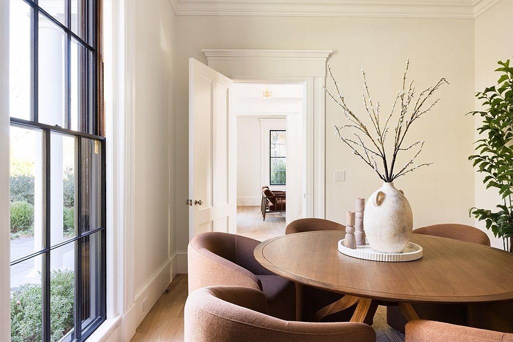 This elegant dining room features a warm, minimalist aesthetic with a round wooden table surrounded by plush, rust-colored upholstered chairs. A large, textured ceramic vase filled with delicate branches serves as a sophisticated centerpiece, while a tall window on the left allows natural light to flood the space. The view through the open doorway reveals a glimpse into an adjacent room, creating a sense of depth and architectural continuity.