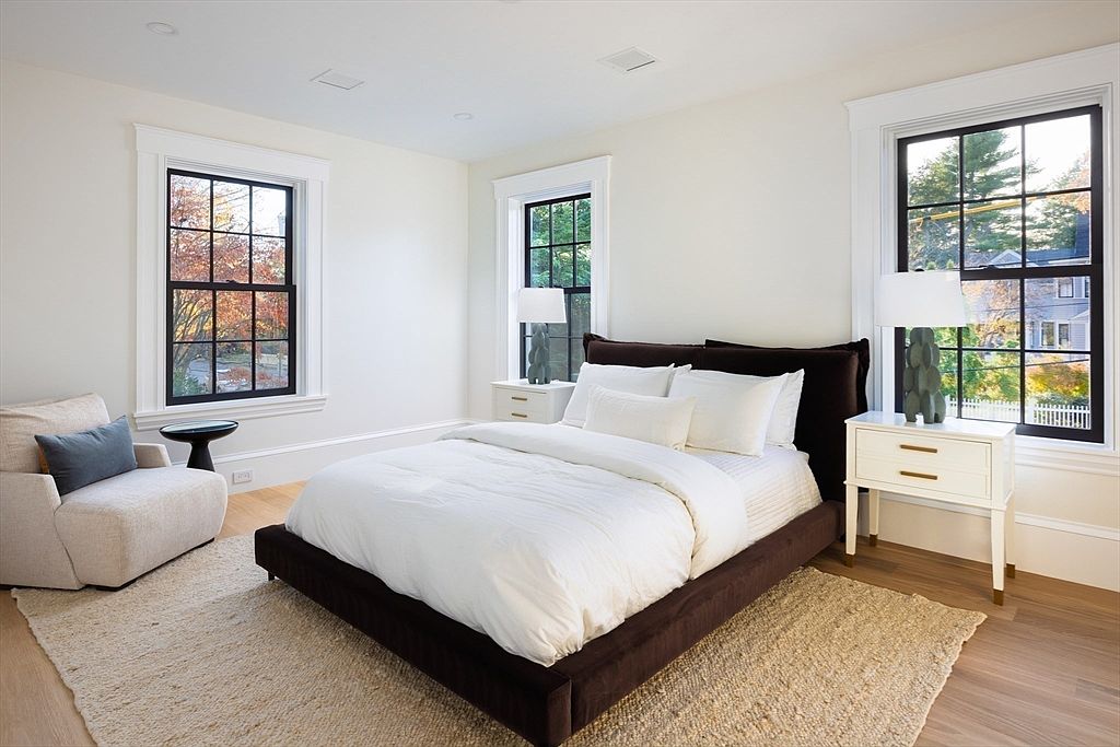 This bright and airy primary bedroom features a plush dark brown upholstered bed frame contrasted by crisp white bedding and a textured jute area rug. The room is framed by three large windows with black mullions that allow for ample natural light, while two matching white nightstands with unique sculptural lamps anchor the space. A cozy armchair with a blue accent pillow sits in the corner, creating a serene and sophisticated retreat.