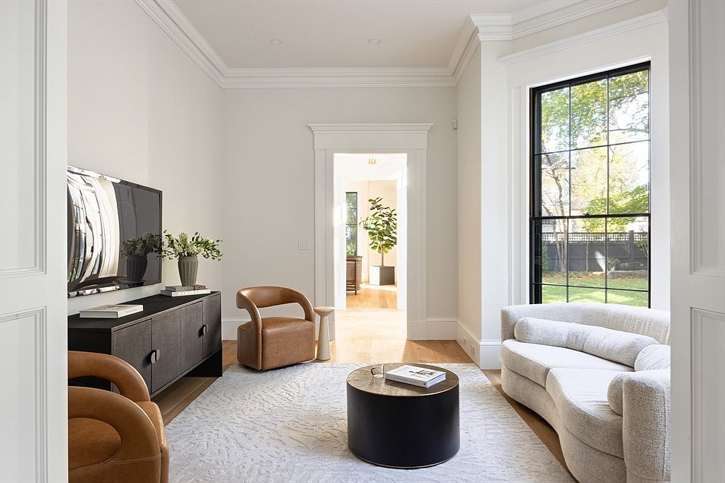 This elegant living room features a sophisticated, neutral color palette with a curved cream sofa, two tan leather armchairs, and a sleek black circular coffee table. A large black-framed window allows natural light to flood the space, while a dark media console sits beneath a wall-mounted television. The room is framed by classic white molding and leads into an adjacent sunlit room, creating an open and airy atmosphere.