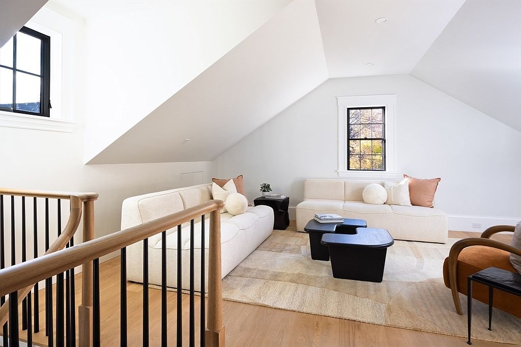 This bright and airy living room is situated in an attic space with sloped ceilings, featuring two cream-colored sofas arranged around a pair of dark, modern coffee tables. The room is anchored by a large, textured area rug and accented with warm-toned throw pillows and a comfortable armchair, creating a cozy yet sophisticated atmosphere. The perspective from the top of the wooden staircase highlights the open, inviting layout and the clean, minimalist aesthetic of the home.