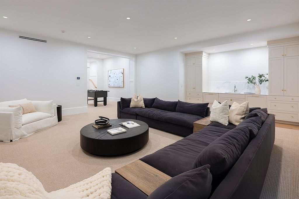 This spacious, modern basement living area features a large, plush dark charcoal sectional sofa arranged around a circular black coffee table, complemented by a separate white armchair. The room is anchored by a built-in wet bar with marble countertops and cabinetry, while a foosball table is visible in the adjacent area, creating a perfect space for entertainment. The neutral color palette, recessed lighting, and clean lines contribute to a sophisticated and inviting atmosphere.