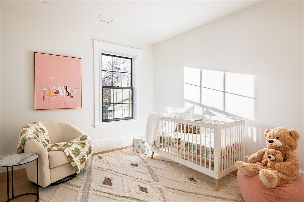 This bright and airy nursery features a white crib, a cozy beige swivel chair with a crochet throw, and a soft area rug with a geometric pattern. A large pink abstract art piece hangs on the wall, while a plush teddy bear sits on a pink beanbag chair, creating a warm and inviting atmosphere. The room is bathed in natural light from a window, highlighting the clean, modern aesthetic perfect for a child's space.