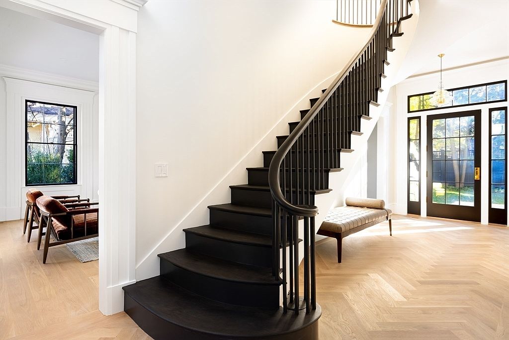 This elegant entryway features a striking curved staircase with dark wood treads and a matching handrail, creating a dramatic focal point against the crisp white walls. The light-toned herringbone hardwood flooring adds warmth and texture, while the large glass-paned front door allows natural light to flood the space. A sleek bench sits beneath the stairs, and a glimpse into an adjacent room reveals a sophisticated seating area, emphasizing the home's open and airy architectural design.