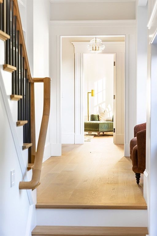This elegant hallway features a staircase with a natural wood handrail and black balusters, leading down to a bright, open transition space. The view extends through a classic doorway into a sunlit room furnished with a green velvet chaise lounge and a modern gold floor lamp. The light-toned hardwood flooring and crisp white trim create a sophisticated, airy atmosphere that emphasizes the home's architectural flow.