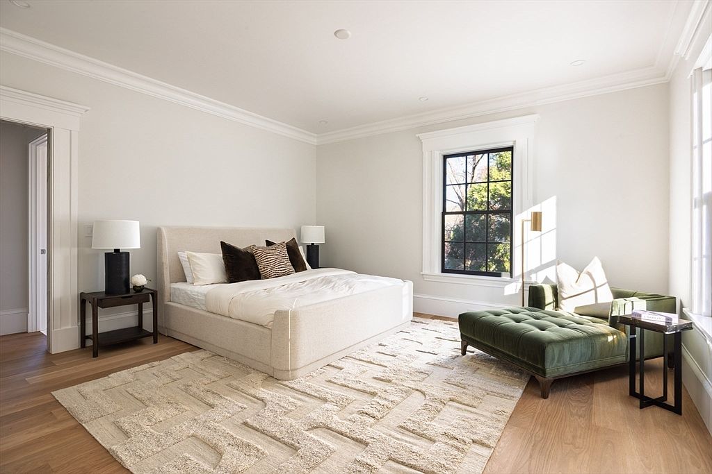 This elegant primary bedroom features a neutral color palette with a plush, light-colored upholstered bed as the focal point, complemented by a textured area rug and warm hardwood flooring. A luxurious green velvet chaise lounge sits near the window, creating a cozy reading nook bathed in natural light. The room is finished with sophisticated black-framed windows and minimalist bedside lamps, offering a serene and high-end atmosphere.