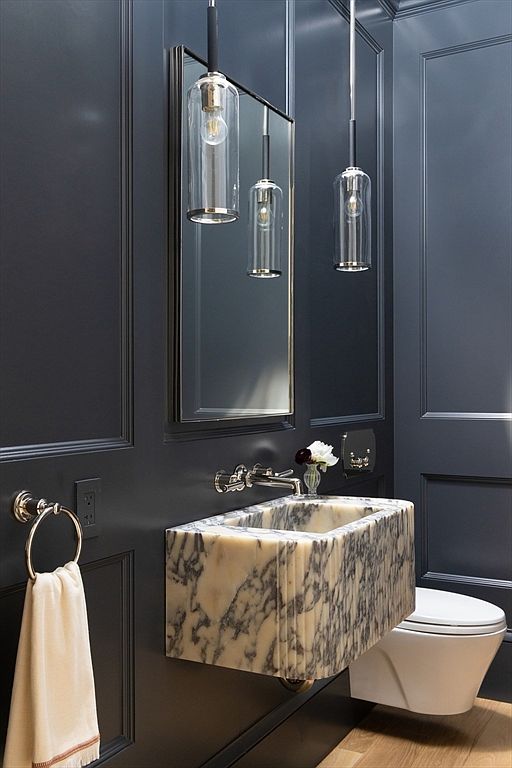 This sophisticated powder room features dramatic dark charcoal wall paneling that provides a striking contrast to the custom, floating marble sink. The space is accented with polished nickel fixtures, including a sleek wall-mounted faucet and modern cylindrical pendant lights hanging before a rectangular mirror. The overall aesthetic is moody, luxurious, and highly curated, making it a standout feature for a high-end home.