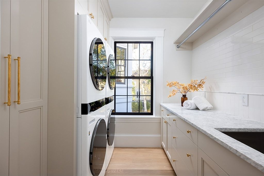 This bright and modern laundry room features a stacked washer and dryer unit set against a clean, neutral color palette. A long marble countertop provides ample workspace, complemented by white subway tile backsplash and elegant gold hardware on the cabinetry. The space is illuminated by natural light streaming through a large, black-framed window, creating a functional yet sophisticated atmosphere.