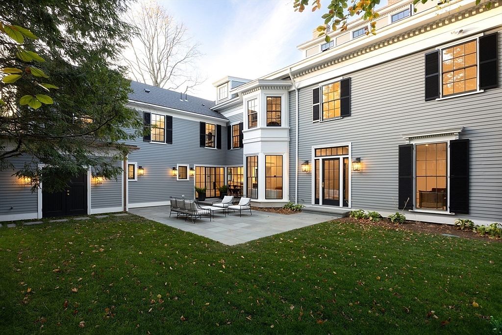 This elegant rear exterior view showcases a classic gray-sided home with a prominent white-trimmed bay window and a stone patio perfect for outdoor seating. The scene is framed by a lush green lawn and mature trees, creating a serene and inviting atmosphere. The architectural design features symmetrical windows with black shutters, highlighting a sophisticated and timeless aesthetic.