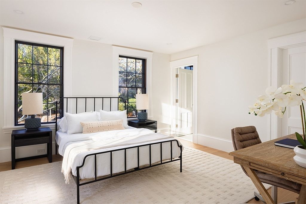 This bright and airy primary bedroom features a classic black metal bed frame centered between two matching nightstands with textured lamps. A wooden desk and chair are positioned in the foreground, creating a functional workspace within the serene sleeping area. The room is finished with crisp white walls, light hardwood floors, and large windows that invite natural light, offering a clean and sophisticated aesthetic.