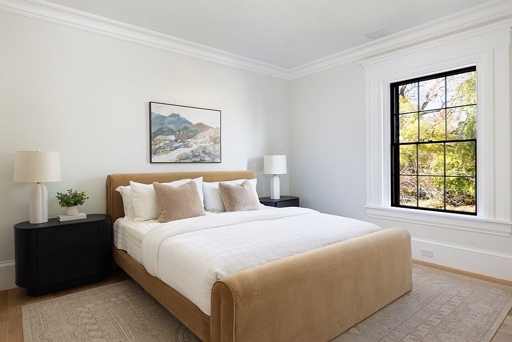 This serene primary bedroom features a plush, camel-colored upholstered bed frame as the focal point, complemented by a neutral color palette and soft textures. The room is anchored by a patterned area rug and flanked by sleek, dark-toned nightstands topped with modern table lamps. A large window with black muntins allows natural light to flood the space, offering a peaceful view of the outdoors.