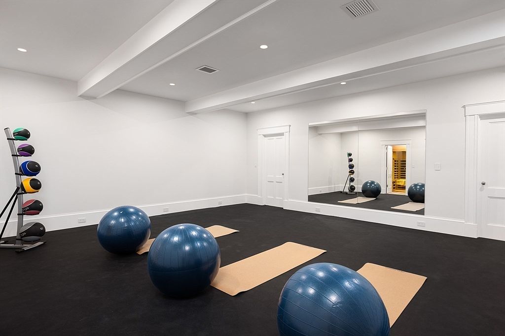 This spacious home gym features a clean, minimalist aesthetic with white walls, high ceilings, and a durable black rubber floor. The room is equipped with a rack of medicine balls, several blue exercise balls, and yoga mats, while a large wall-mounted mirror enhances the sense of space and reflects a glimpse of an adjacent sauna. The perspective is wide and inviting, showcasing a professional-grade workout area designed for fitness and wellness.