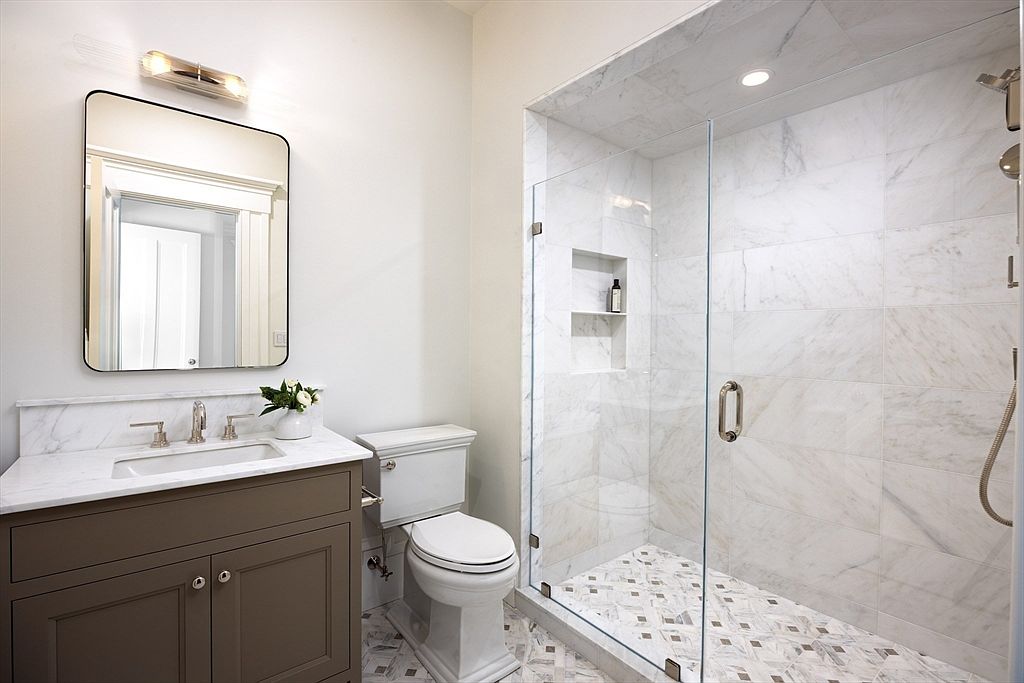 This elegant primary bathroom features a sleek vanity with a marble countertop, a modern rectangular mirror, and a spacious walk-in shower enclosed by glass. The shower is beautifully tiled with white marble and includes a built-in niche, while the floor showcases a sophisticated patterned tile design. The overall aesthetic is clean, bright, and luxurious, offering a spa-like atmosphere.