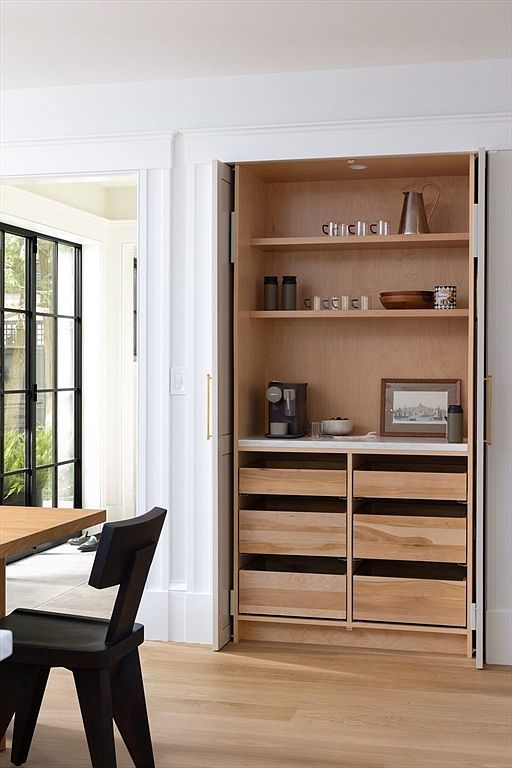 This image showcases a sophisticated, built-in pantry or coffee station featuring light wood cabinetry and open shelving. The unit is integrated into a white-trimmed wall, offering a clean, minimalist aesthetic with a coffee machine, decorative items, and organized drawers. A modern black chair sits in the foreground, providing a glimpse into an adjacent room with large glass-paned doors.