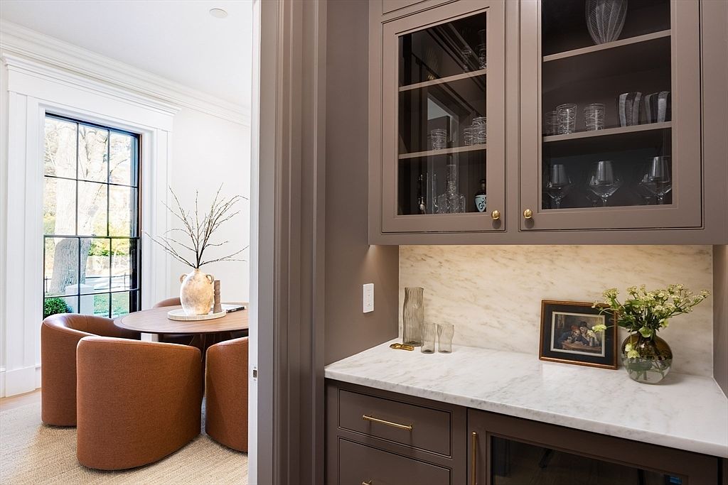 This image features a sophisticated butler's pantry or wet bar area with custom taupe cabinetry, marble countertops, and a marble backsplash. To the left, a doorway opens into a bright dining area furnished with a round wooden table and modern, curved rust-colored chairs. The composition creates a seamless transition between the functional bar space and the elegant dining room, highlighting a refined, high-end interior design aesthetic.