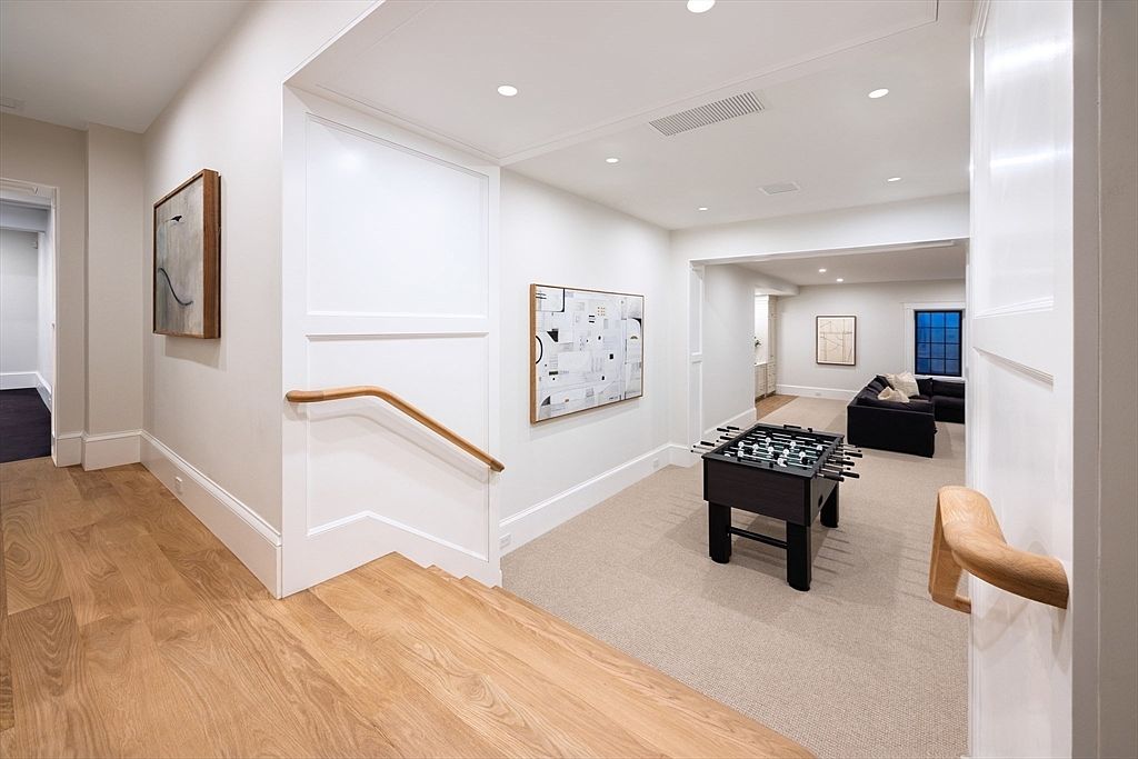 This bright, modern hallway features light oak hardwood flooring that transitions into a plush, neutral-toned carpeted area. A foosball table serves as a focal point in the open space, which leads into a cozy living area with a dark sofa and minimalist artwork. The clean white walls, recessed lighting, and natural wood handrails create a sophisticated and inviting atmosphere.