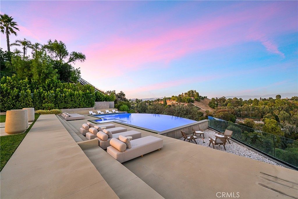 This stunning infinity-edge pool overlooks a lush, rolling landscape under a vibrant sunset sky. The expansive concrete deck is furnished with multiple modern lounge chairs, creating a luxurious outdoor living space perfect for relaxation and entertaining. The sleek, minimalist design emphasizes the seamless transition between the pool and the natural scenery beyond.