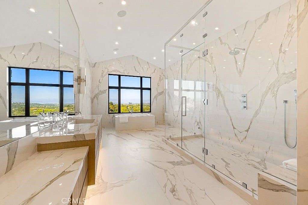 This luxurious primary bathroom features floor-to-ceiling marble tiling, a spacious walk-in glass shower, and a deep soaking tub positioned beneath a large window with scenic views. The double vanity with integrated sinks and warm under-cabinet lighting adds a sophisticated, modern touch to the bright, airy space. The perspective is wide-angle, capturing the expansive layout and high-end finishes that define this elegant retreat.