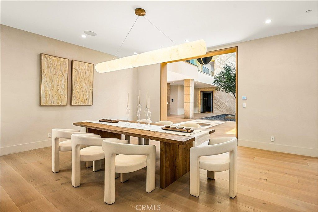 This elegant dining room features a long, rustic wooden table surrounded by modern, cream-colored curved chairs, all illuminated by a striking, elongated cylindrical pendant light. The room boasts light-toned hardwood floors and minimalist decor, with a seamless transition through a large open doorway to an outdoor courtyard area. The overall aesthetic is sophisticated, airy, and contemporary, emphasizing a clean and inviting atmosphere.