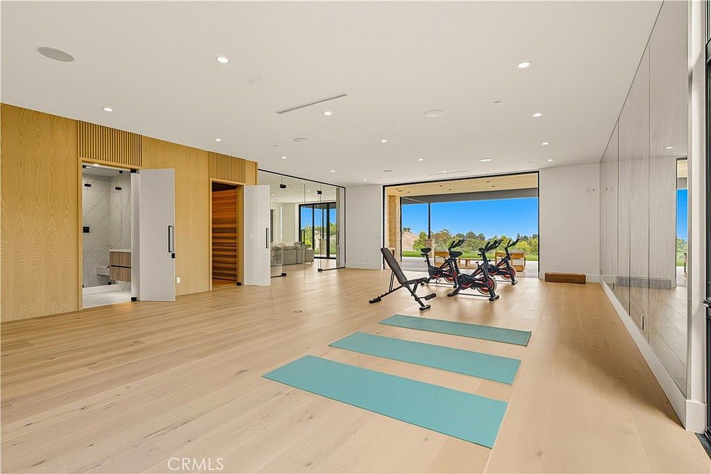 This spacious, modern home gym features light-toned hardwood flooring and a minimalist aesthetic, highlighted by a wall of floor-to-ceiling mirrors. The room is equipped with multiple stationary exercise bikes and a weight bench, with large glass doors opening to a scenic outdoor view. The clean lines and bright, airy atmosphere create an inviting and professional workout environment.