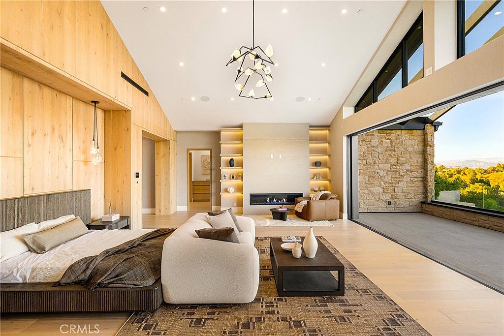 This expansive primary bedroom features a sophisticated, modern design with high vaulted ceilings and warm wood-paneled walls. The room is anchored by a plush, low-profile bed and a cozy seating area centered around a sleek, linear fireplace, all opening onto a spacious balcony with panoramic views. The neutral color palette and minimalist aesthetic create a serene, luxurious atmosphere perfect for a high-end residence.