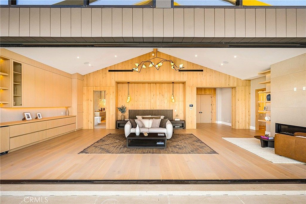 This expansive primary bedroom suite features a warm, modern aesthetic with light wood paneling and vaulted ceilings. The room is anchored by a central seating area with a plush white sofa and a dark area rug, while a fireplace and built-in shelving add functional elegance. The perspective is captured from an open-air transition space, emphasizing the seamless flow between the indoor living area and the outdoors.