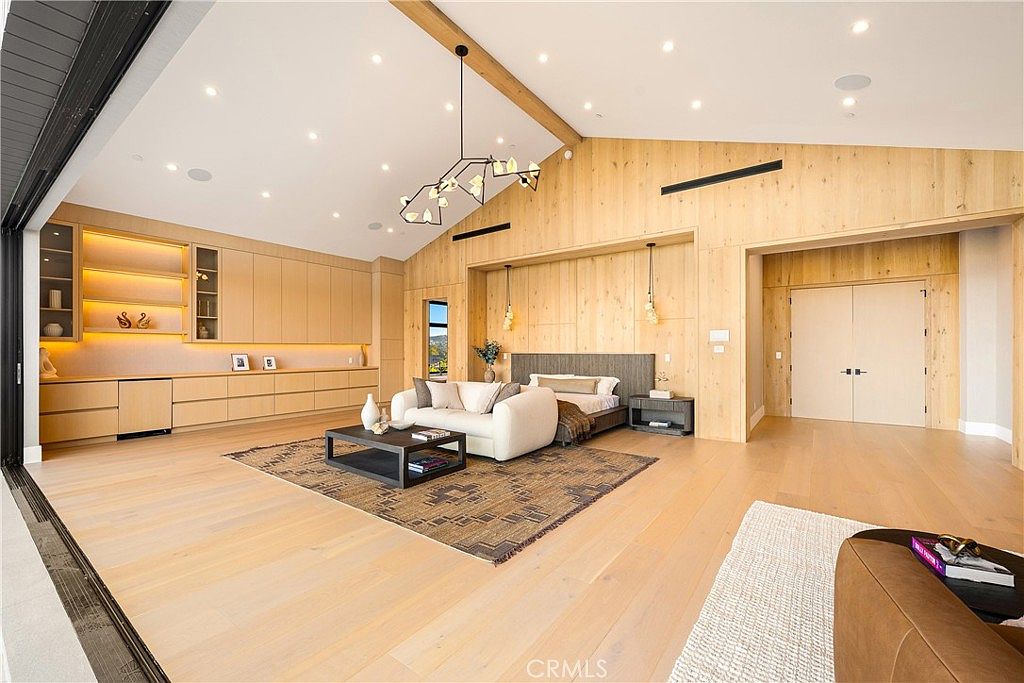 This expansive primary bedroom features a sophisticated, modern aesthetic with light wood paneling, vaulted ceilings, and recessed lighting. The room is anchored by a plush white sofa on a patterned area rug, with a bed positioned against a wood-paneled accent wall, all opening up to an outdoor space through a large sliding glass door. The overall impression is one of luxurious, airy comfort with high-end finishes and a seamless indoor-outdoor flow.