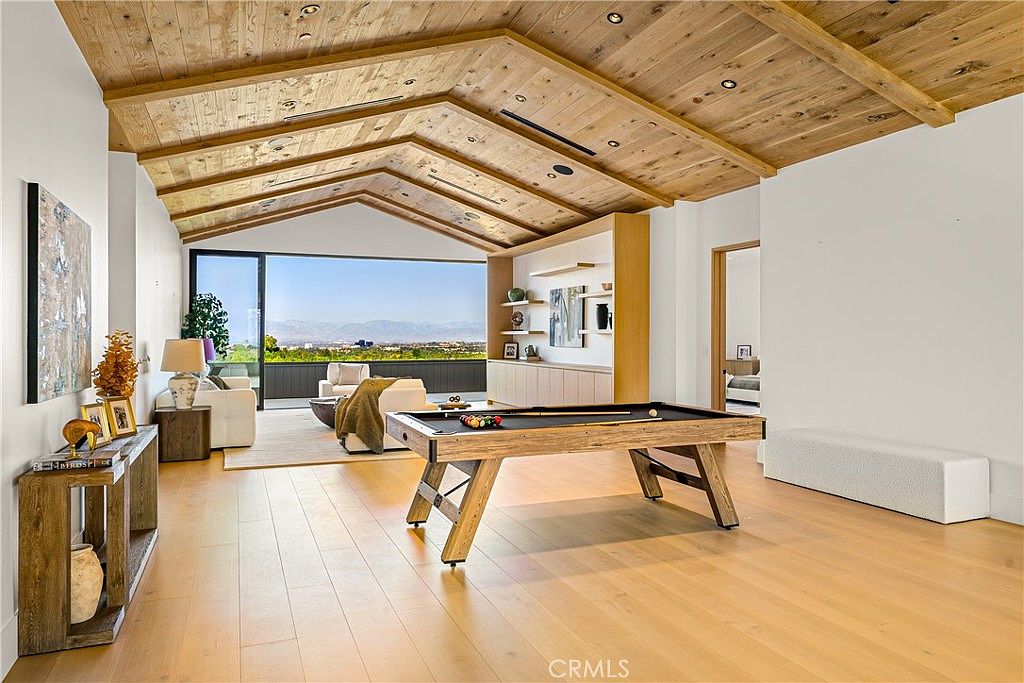 This spacious, open-concept living area features a striking vaulted wood-paneled ceiling and light-toned hardwood flooring, creating a warm and modern aesthetic. The room is anchored by a central pool table and flows seamlessly toward a large glass wall that reveals a panoramic view of the distant mountains. A built-in shelving unit and minimalist decor enhance the sophisticated, airy atmosphere of this high-end living space.