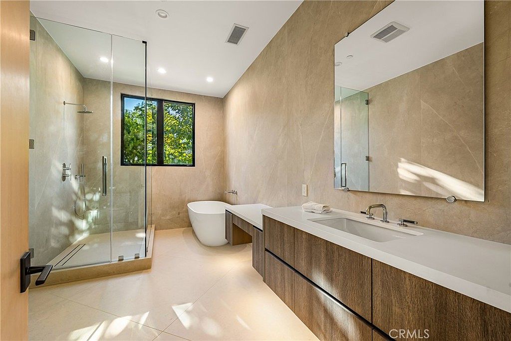This modern primary bathroom features a sleek, minimalist design with large-format beige stone tiles covering the walls and floor. The space includes a floating wood-grain vanity with a white countertop, a frameless glass-enclosed walk-in shower, and a contemporary freestanding soaking tub positioned near a window. The perspective is from the doorway, capturing the clean lines and high-end finishes that create a spa-like atmosphere.