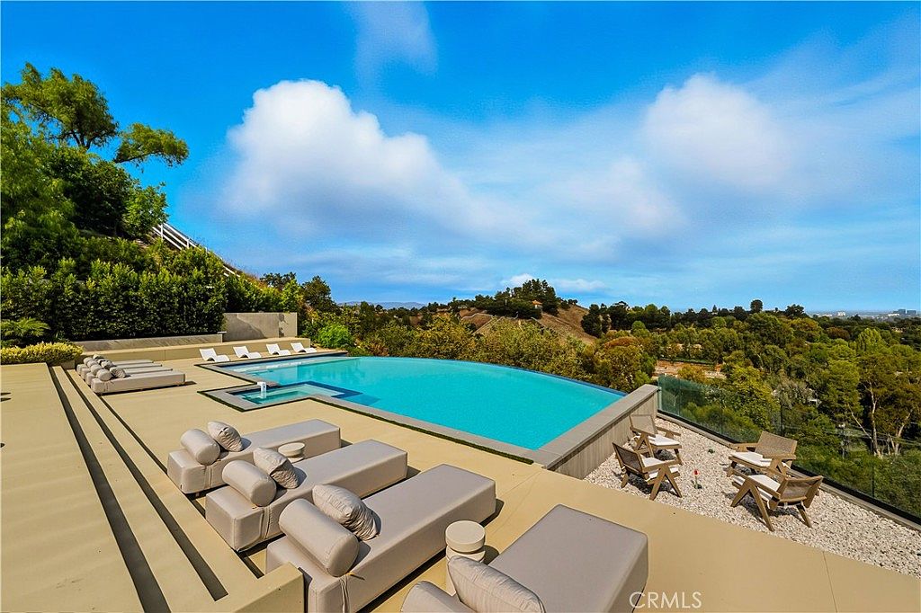 This stunning outdoor space features a sleek, infinity-edge swimming pool that seamlessly blends with the expansive hillside views. The surrounding deck is furnished with multiple modern lounge chairs, creating a luxurious and serene atmosphere perfect for relaxation. The elevated perspective captures the clean lines of the pool design against the backdrop of lush greenery and a bright, clear sky.
