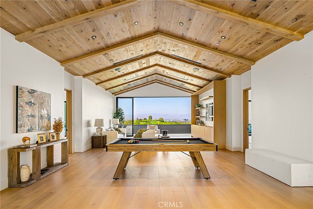 This spacious, modern living area features a striking vaulted ceiling with exposed wooden beams and light-toned hardwood flooring. A central pool table serves as the focal point, while the rear wall consists of expansive floor-to-ceiling glass that frames a scenic outdoor view. The room is elegantly furnished with a minimalist aesthetic, including built-in shelving and a long, low-profile bench, creating a sophisticated and airy atmosphere.
