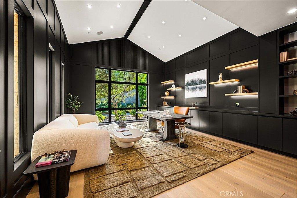 This sophisticated home office features dramatic black paneled walls, a vaulted ceiling, and a large window that floods the space with natural light. The room is furnished with a modern desk, a plush cream-colored sofa, and a textured area rug, creating a luxurious and professional atmosphere. The clean lines and high-contrast color palette provide a contemporary and refined aesthetic suitable for a high-end residence.