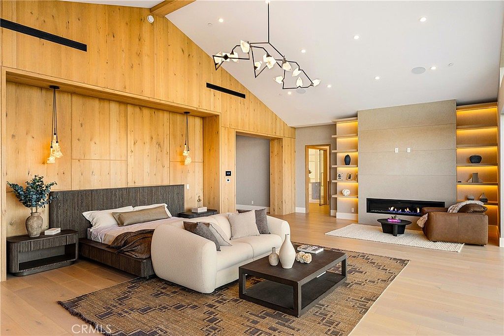 This expansive primary bedroom features a sophisticated blend of warm wood paneling and modern design elements, including a vaulted ceiling and a striking geometric chandelier. The room is anchored by a plush, low-profile bed and a cozy seating area centered around a sleek, linear fireplace built into a minimalist wall. The open-concept layout creates a luxurious, airy atmosphere, perfect for high-end residential living.