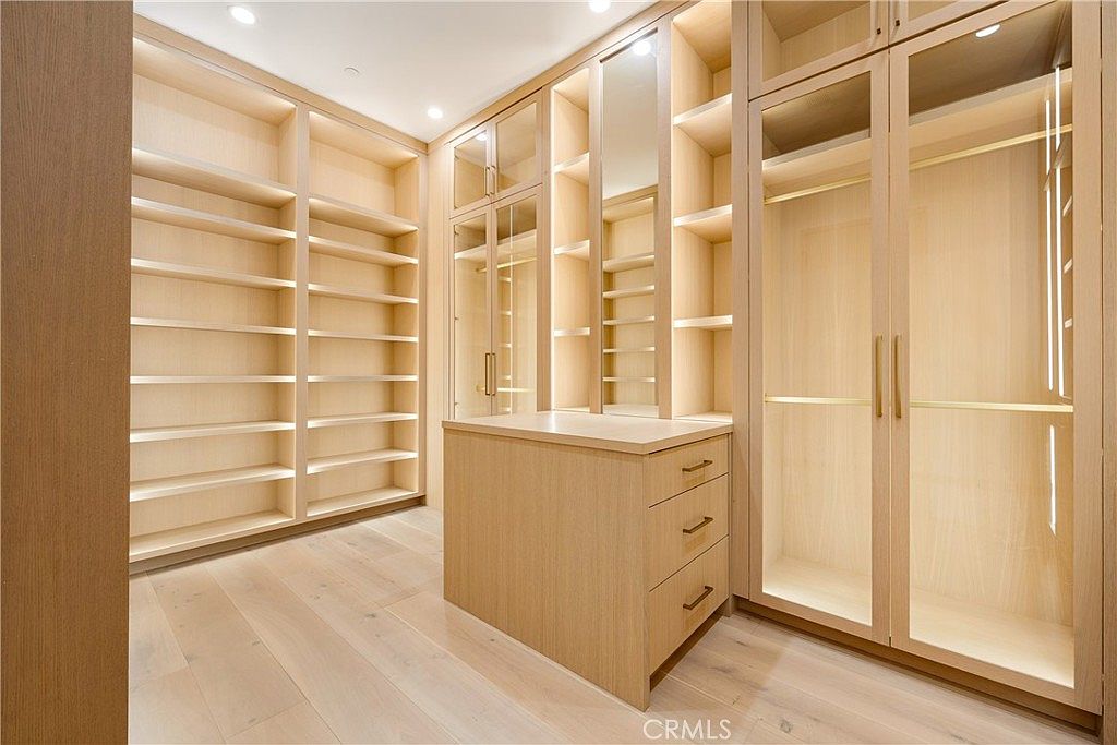 This luxurious walk-in closet features custom light-oak cabinetry, providing an abundance of open shelving, hanging space, and a central island with three drawers. The design includes glass-fronted cabinet doors and a mirrored panel, creating a bright, sophisticated, and organized atmosphere. The light wood flooring and recessed lighting enhance the clean, modern aesthetic of this high-end storage space.