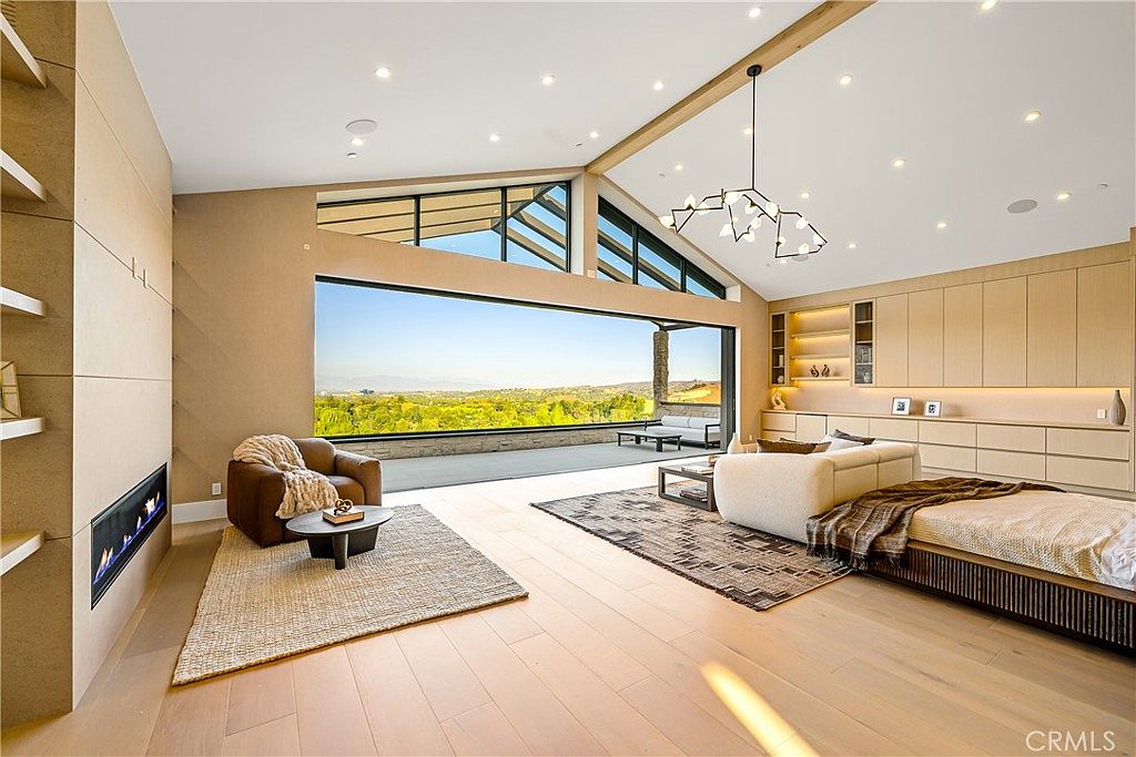 This expansive, modern living room features a seamless indoor-outdoor flow, highlighted by a massive floor-to-ceiling glass wall that opens onto a scenic patio with panoramic hillside views. The space is anchored by a sleek, linear fireplace built into a light-toned wood wall, complemented by a contemporary chandelier and minimalist cabinetry. The open-concept design, warm wood flooring, and neutral color palette create an airy, sophisticated atmosphere perfect for luxury living.