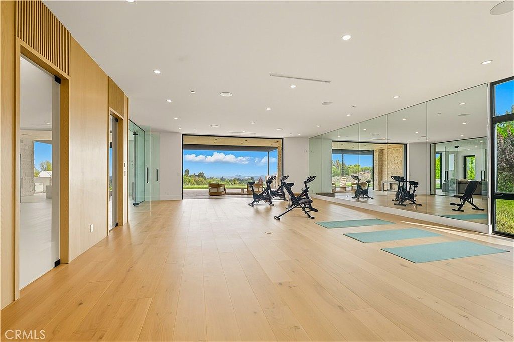 This spacious, modern home gym features light hardwood flooring, floor-to-ceiling mirrored walls, and large sliding glass doors that open to a scenic outdoor view. The room is equipped with multiple stationary exercise bikes and yoga mats, creating a bright and airy atmosphere perfect for fitness. The minimalist design and natural light contribute to a luxurious and inviting workout environment.