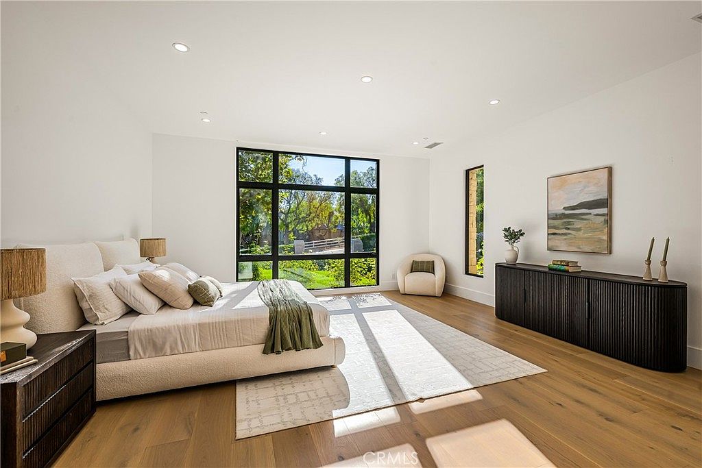 This spacious and serene primary bedroom features light oak hardwood flooring, crisp white walls, and a large, modern window grid that floods the room with natural light. The room is anchored by a plush, cream-colored upholstered bed and a sleek, dark wood sideboard, creating a sophisticated and minimalist aesthetic. The perspective is wide-angle, capturing the airy, tranquil atmosphere of this high-end residential suite.