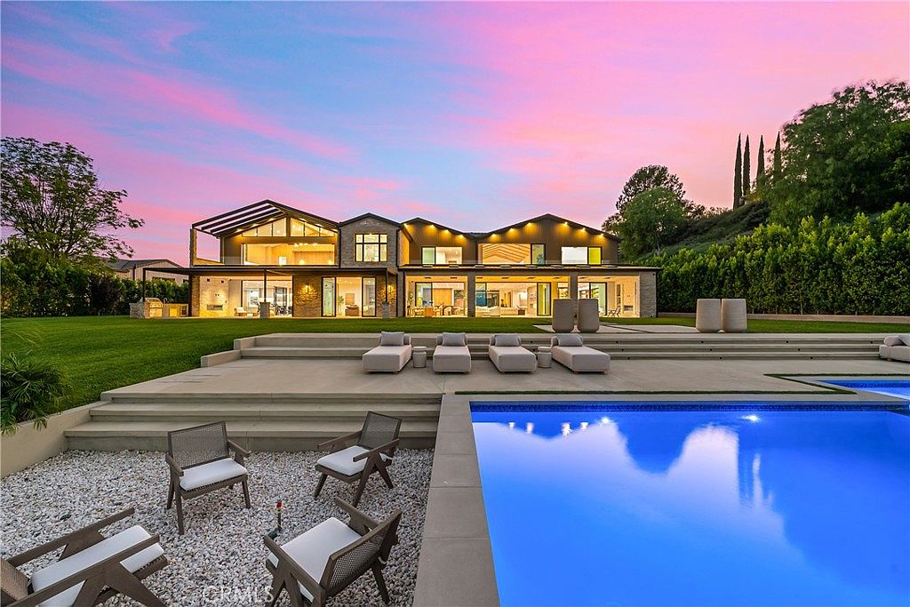 This stunning rear exterior view showcases a modern luxury estate at sunset, featuring a sprawling lawn, a large infinity-edge pool, and a multi-level patio area. The home boasts expansive glass walls that seamlessly blend indoor and outdoor living, complemented by warm architectural lighting. The scene is framed by lush greenery and a vibrant, colorful sky, creating an inviting and sophisticated atmosphere.