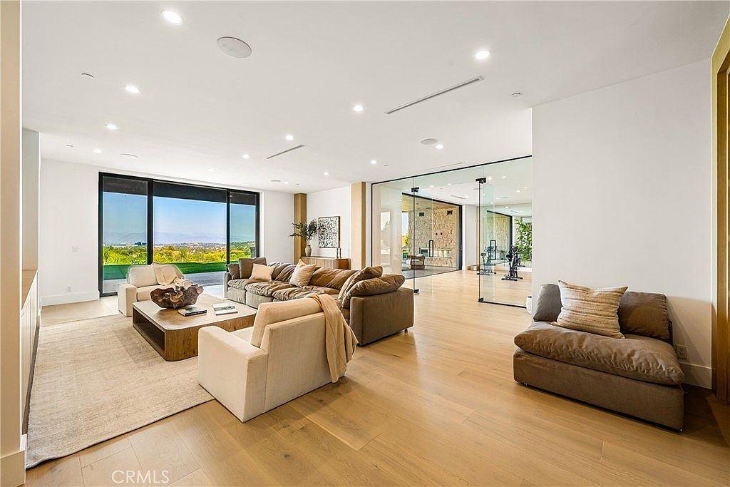 This expansive, modern living room features an open-concept design with light hardwood flooring and floor-to-ceiling glass walls that offer panoramic views of the landscape. The space is furnished with a large, plush sectional sofa, a contemporary coffee table, and a separate lounge chair, all arranged to emphasize the airy, minimalist aesthetic. A glass-enclosed home gym is visible in the background, adding to the sophisticated and high-end feel of the residence.