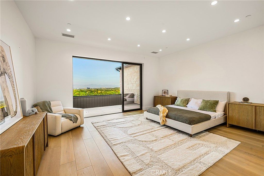 This spacious primary bedroom features a contemporary design with light hardwood flooring, a neutral color palette, and a large sliding glass door that opens onto a private balcony with scenic views. The room is anchored by a modern upholstered bed, a textured area rug, and minimalist wooden cabinetry, creating a serene and sophisticated atmosphere. The perspective is wide-angle, capturing the seamless transition between the luxurious indoor living space and the expansive outdoor landscape.