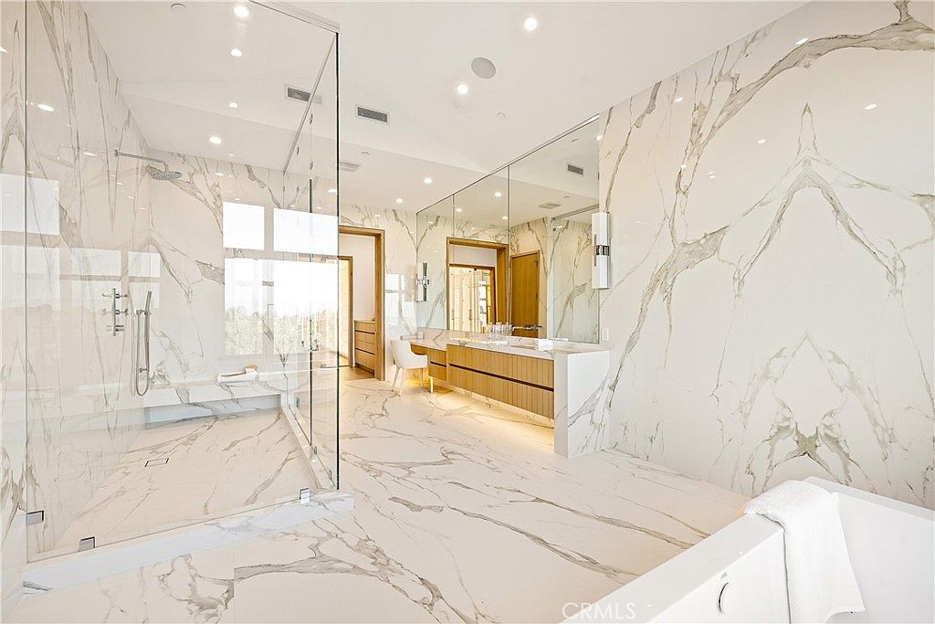 This luxurious primary bathroom features floor-to-ceiling white marble with dramatic gold veining, creating a sophisticated and high-end aesthetic. The space includes a spacious walk-in glass shower, a floating wooden vanity with dual sinks, and a deep soaking tub in the foreground. The bright, open layout is enhanced by large mirrors and recessed lighting, offering a clean and modern spa-like atmosphere.
