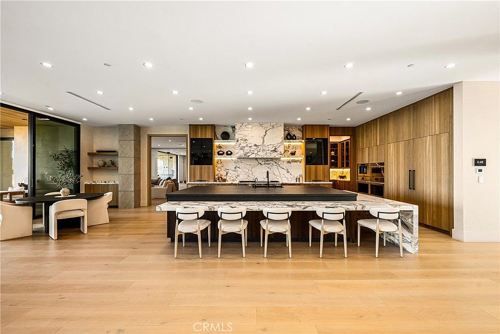 This expansive, modern kitchen features a massive central island with a striking marble waterfall countertop and seating for four. The space is defined by high-end finishes, including warm wood cabinetry, integrated appliances, and a dramatic marble backsplash that serves as a focal point. The open-concept design seamlessly flows into a dining area, creating a sophisticated and airy atmosphere perfect for luxury living.